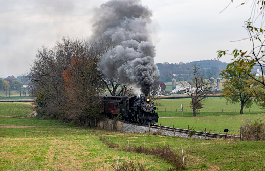 Ronks, Pennsylvania, November 4, 2023 - View of an Antique Steam Passenger Train Approaching Coming Out of a Group of Trees, Blowing Smoke on a Fall Day