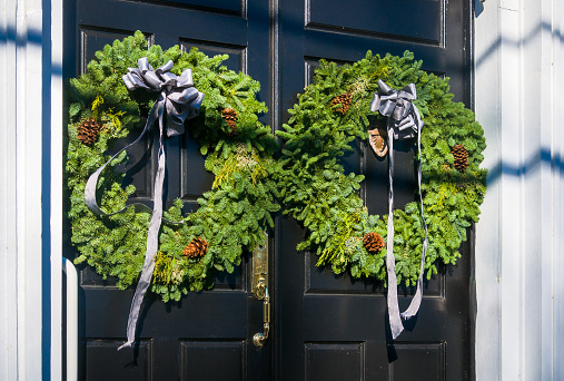 Two Christmas wreaths made of greens, pinecones and other natural adornments are decorated with gray ribbons on a set of black double doors