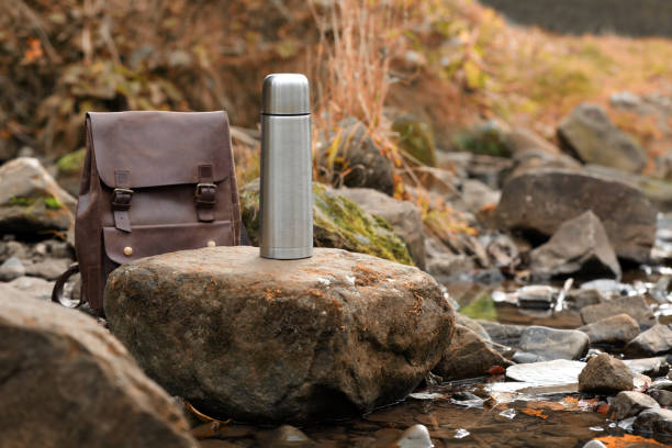 istock Metallic thermos and leather backpack on stone near river, space for text 1858198857