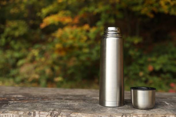istock Metallic thermos and cup lid on wooden bench outdoors, space for text 1858199888