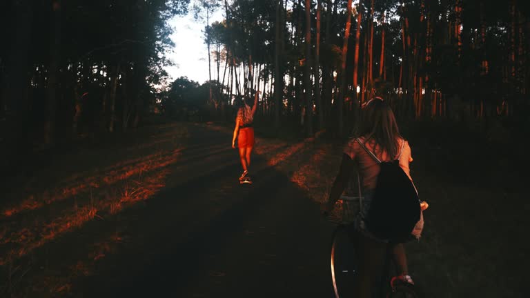 Silhouette of two women riding bicycle and skateboard on forest road