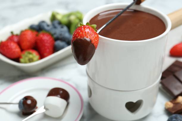 istock Fondue pot of melted chocolate and fork with strawberry on white marble table, closeup 1858219482