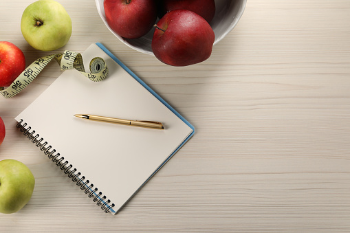 Healthy diet. Fresh apples, measuring tape, notebook and pen on light wooden table, flat lay. Space for text