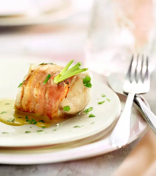 monkfish filet wraped in bacon served with a sauce and a garnish of parsley, on a white plate brightly lit