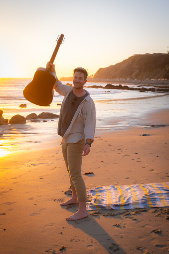 A handsome man stands on beach at sunset and holds his guitar into the air.