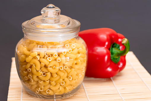 Uncooked Chifferi Rigati Pasta in Glass Jar with Red Bell Pepper on Bamboo Mat