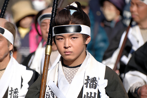 A youthful individual, dressed as a samurai and holding a spear, marching in the procession at the Yamashina Gishi Festival. This annual event takes place every December 14th in a distinguished ward of Kyoto and is dedicated to the celebrated story of the 47 ronin. It serves as a historical re-enactment, paying homage to these samurai who became without a leader after their master's forced suicide due to a conflict with a higher-ranking official. The festival, featuring a parade through the ward, effectively dramatizes the samurai's pursuit of vengeance thereafter.