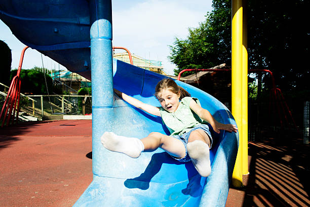 A young girl enjoys playful moments on a bright blue playground slide at a neighborhood park. Autumn leaves and warm light create a cheerful, carefree day.