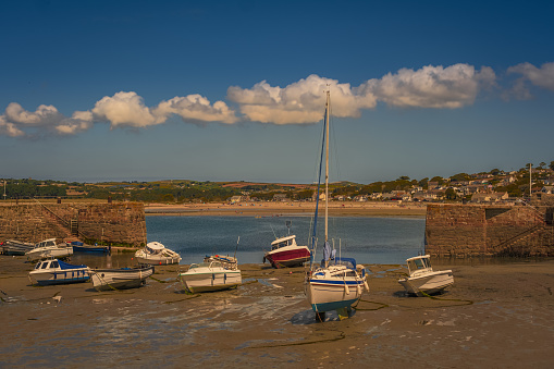 View of boats sitting on sand in low tide in Cornwall, England, with distant view of coastal town across bay and blue sky with puffy clouds in background in summer