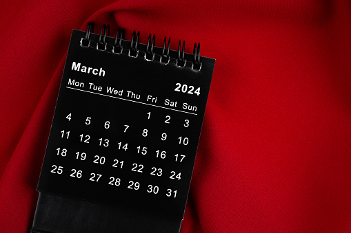 March 2024 Black spiral desktop calendar on red textile.
