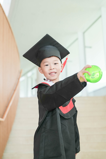 Asian elementary age boys wearing graduation caps and gowns smile while standing inside school