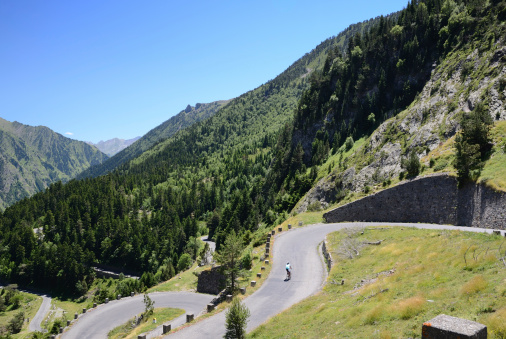 There is a green slope with a serpentine road in summer mountains. A cyclist is pedaling upwards.