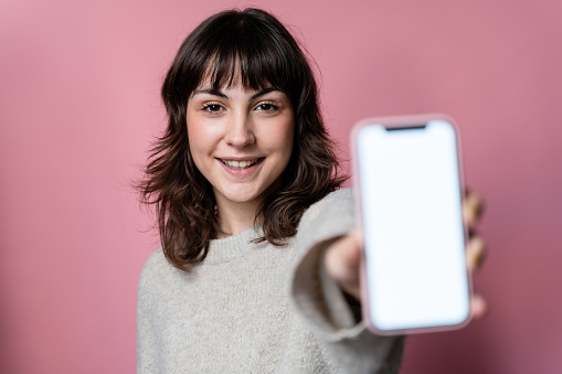 Portrait of a beautiful young woman showing a smartphone on a pink background.