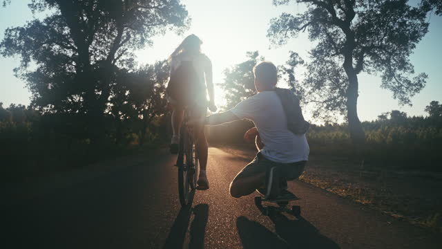 SLO MO Silhouette of woman riding bicycle pulling her male friend skateboarding on countryside road