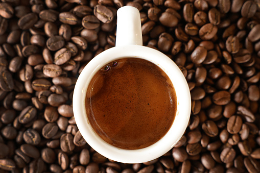 A close up view from directly above a clean white espresso cup filled with a fresh coffee shot. Background of freshly roasted coffee beans.