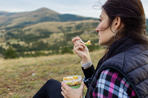 Hiker woman with backpack is taking a break and eating healthy meal, fruit and cereal after walking in nature at cloudy autumn day. Healthy lifestyle outdoors.