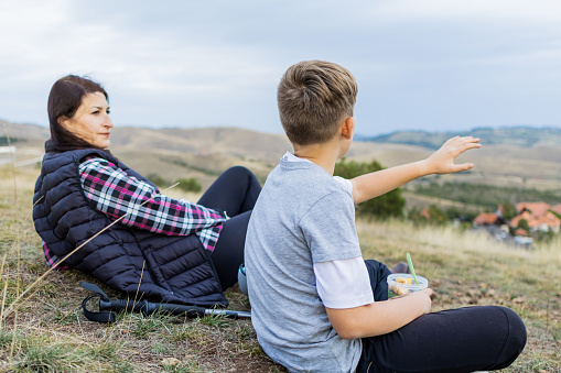 Hikers, mother and her son with backpacks are taking a break and eating healthy meal, fruit and cereal after walking in nature at cloudy autumn day. Healthy lifestyle outdoors.