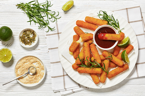 fried breaded mozzarella sticks served with dipping sauce, lime slices and fresh rosemary on white platter on white wooden table with ingredients, horizontal view from above, flat lay