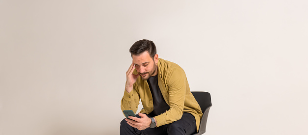 Stressed businessman reading messages on smart phone while sitting on chair over white background