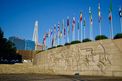 Seoul, South Korea - June 2, 2023: Intricate figures carved on a wall next to Mongchon Lake in Olympic Park, with flagpoles atop and Lotte World Tower in the distance.