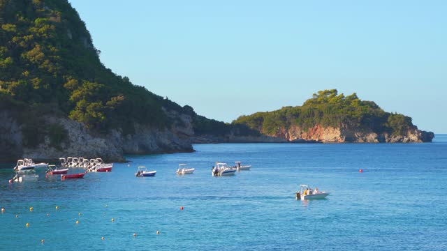 Afternoon sun shines to calm blue sea at Liapades bay, docked boats moving slowly in distance, rocky cliffs with trees background