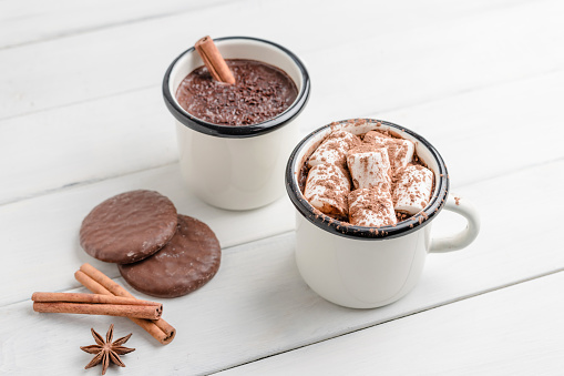Homemade spicy hot chocolate drink with marshmallows in enamel cup on white wooden table with cinnamon stick, cookies and star anise.