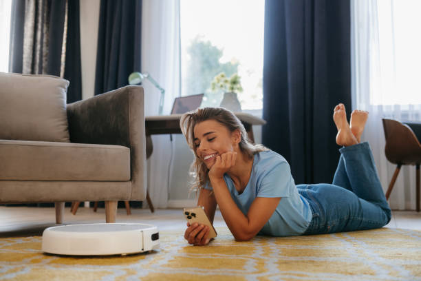 istock Happy young woman lying down on the floor and using mobile phone to control smart vacuum cleaner robot 1859598201