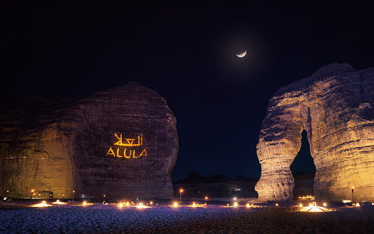 Jabal AlFil - Elephant Rock in evening, landscape illuminated, text Alula and Arabic translation projected to stone wall seats for people set up on the ground, crescent moon - composite image - above