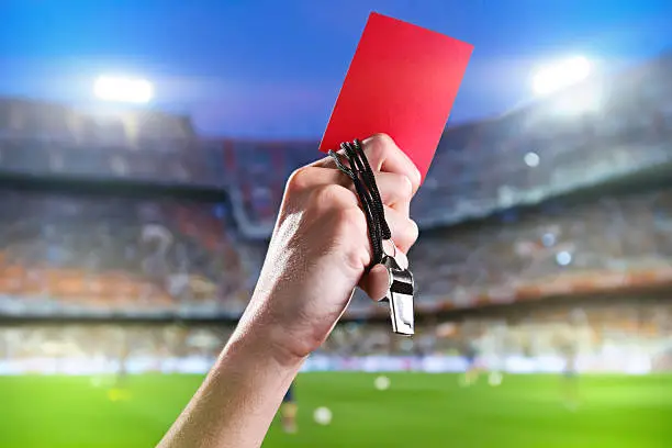Hand of referee with red card and whistle in the soccer stadium.