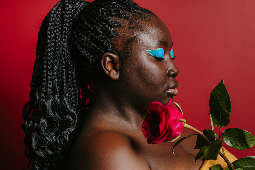 Beautiful plus size African woman with beautiful make-up holding single rose near face on red background