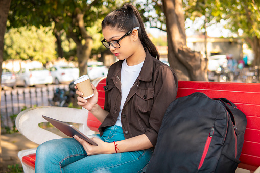 Portrait of beautiful Asian/Indian young woman sitting relaxed on a bench in park and using a digital tablet while having a cup of coffee in the outdoor fresh air at day time.