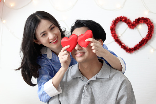 Happy Asian couple spending time together at home. Beautiful smiling woman girl holding red heart to cover her boyfriend eyes. Romantic lover celebrating anniversary on Valentine Day.