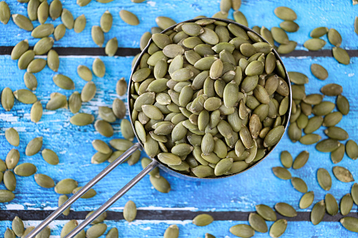 Stock photo showing close-up, elevated view of some pumpkin seeds, also known as pepita, piled high in a handled, stainless steel measuring cup against a woodgrain background. Raw pumpkin seeds are considered to be a very healthy snack food and are high in vitamin K, manganese, antioxidants and protein, boasting a list of health benefits and may aid bladder health.