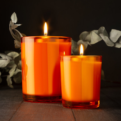 Atmosphere burning orange candles in orange glass with dry leaves as elegant home decor on black wood table in darkness, copy space. Romantic relaxation background with candles for text, advertising.