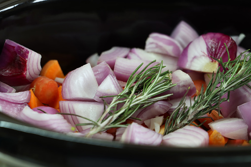 Stock photo showing close-up, elevated view of slow cooker containing chopped ingredients including vegetables and fresh herbs ready to be cooked.
