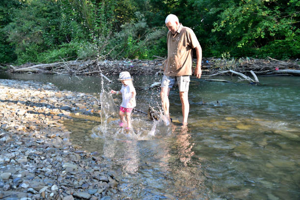 Grandfather and granddaughter are playing together by the river, throwing stones into the water stock photo