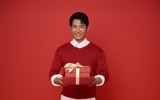 Young man of Asian ethnicity wear red sweater hold red present box with gift ribbon bow isolated on red background. birthday, valentines day, new year concept.