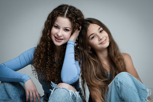 Studio portrait of two beautiful smiling sisters in blue shirts and jeans against gray background