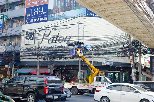 Thai worker is repairing stoplight. Man is in basket of small semitruck crane above ruling traffic. in background are buildings. In top foreground is footbrudge. Scene is at junction of Sutthisan Rd and Ratchadaphisek Rd near BTS station Sutthisan