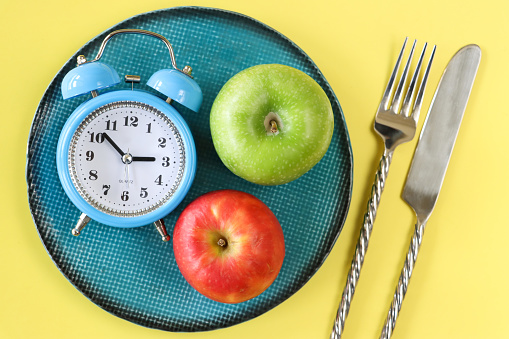 Stock photo showing close-up, elevated view of a healthy eating and intermittent fasting diet concept depicted by a plate containing fruit and double bell alarm clock.