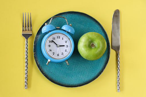 Stock photo showing close-up, elevated view of a healthy eating and intermittent fasting diet concept depicted by a plate containing fruit and double bell alarm clock.