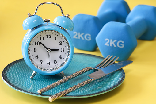 Stock photo showing close-up view of a healthy eating and intermittent fasting diet concept depicted by a plate containing a double bell alarm clock and stainless steel knife and fork besides a pair of hand weights.