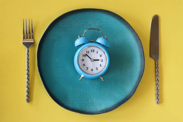 Image of blue metal case, retro alarm clock, double bell, analogue display clock with white dial on turquoise plate, stainless steel knife and fork, yellow background, elevated view