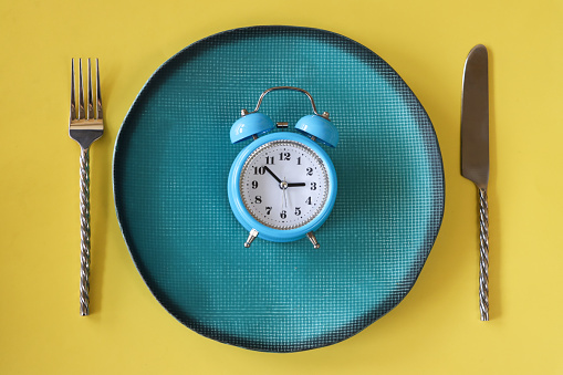 Stock photo showing close-up, elevated view of a healthy eating and intermittent fasting diet concept depicted by a plate containing a double bell alarm clock.