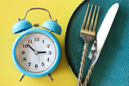 Stock photo showing close-up, elevated view of a healthy eating and intermittent fasting diet concept depicted by a plate containing stainless steel knife and fork besides a double bell alarm clock.