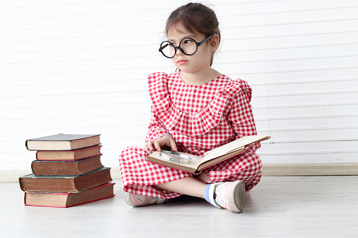 Serious concentrate Asian girl wearing glasses while sitting on floor with book stack in white wall room. Cute child with pile of books, kid education, learning, reading and exploring discovery.