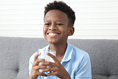 Happy smiling African boy drinking fresh milk from a glass while sitting on the sofa in the living room. children grow up with eating healthy food, protein and calcium rich food are essential for kid