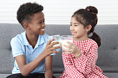 Happy smiling African boy and Asian girl drinking fresh milk together while sitting on sofa in living room. children grow up with eating healthy food, protein calcium rich food are essential for kid.