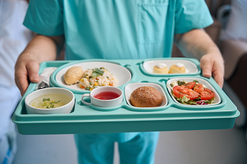 Cropped photo of nursing assistant holding meal tray in hands while standing in hospital room