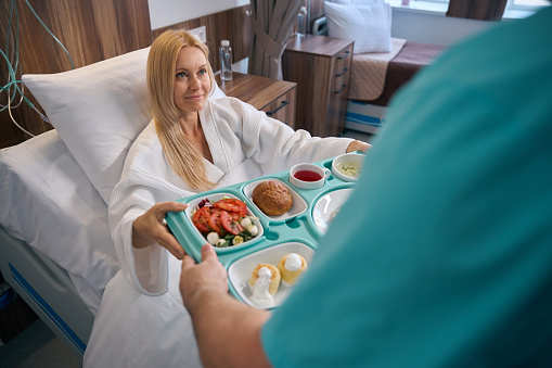 Smiling woman seated in hospital bed taking meal tray from nurse hands